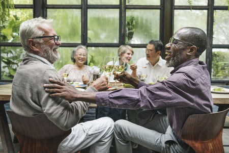 Group of seniors caround a dinner table, two men in the foreground are clinking glasses 